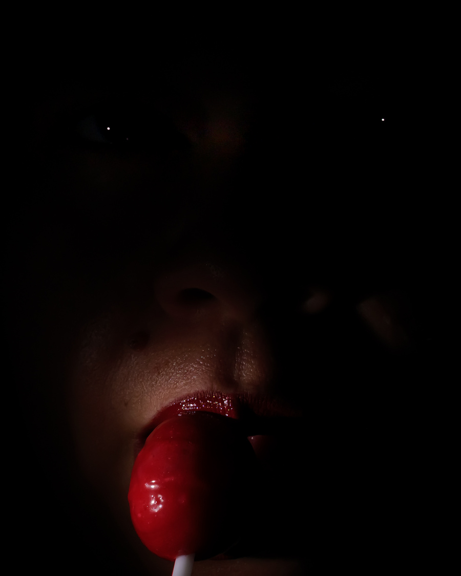 Primer plano cinematográfico de un rostro en sombras, con labios rojos y una chupeta roja frente a la boca.